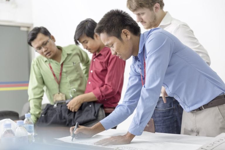 Four men bend over a table on which one of them is writing something