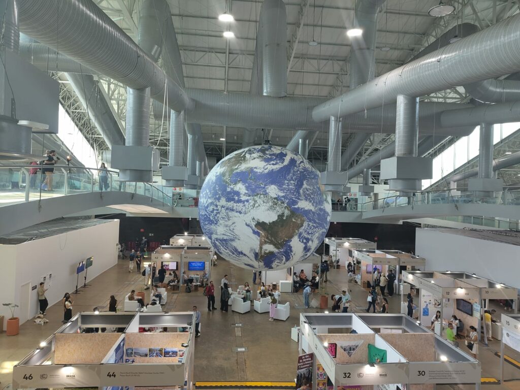 Inside the COP30 building, a large model of the earth can be seen in the background. People are walking between stands in the hall.