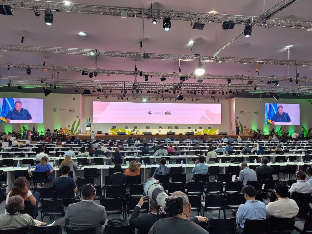 In the front of a big room filled with rows of tables and people, there is a screening of a man talking on the stage.