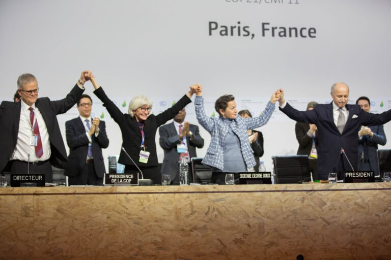 At COP21 the director, president, president of the COP and the executive secretary of UNFCCC are holding their hands up in celebration on a stage.