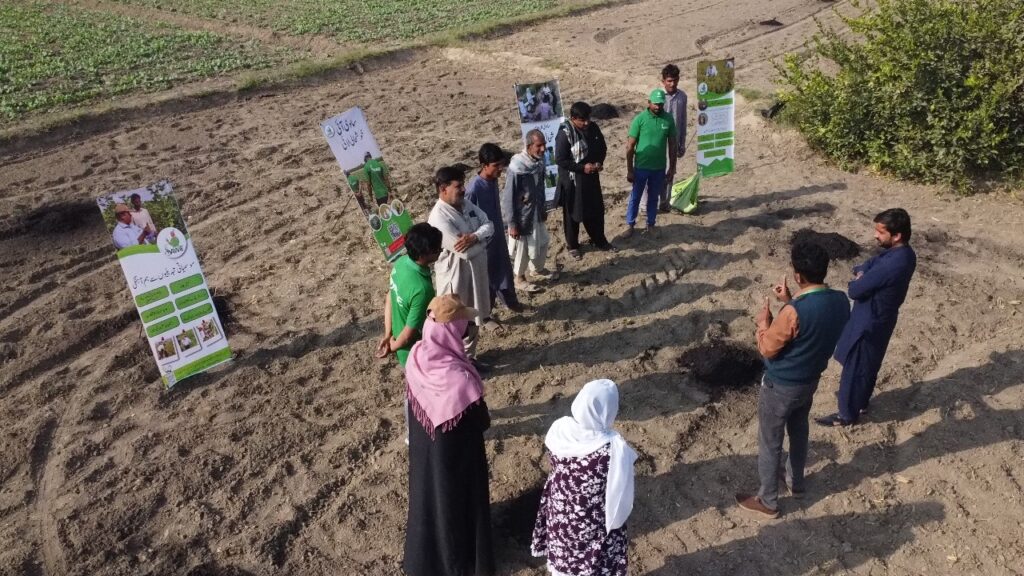 A group of men and women standing in a semi-circle on a bare agricultural field, listening to a person giving instructions.